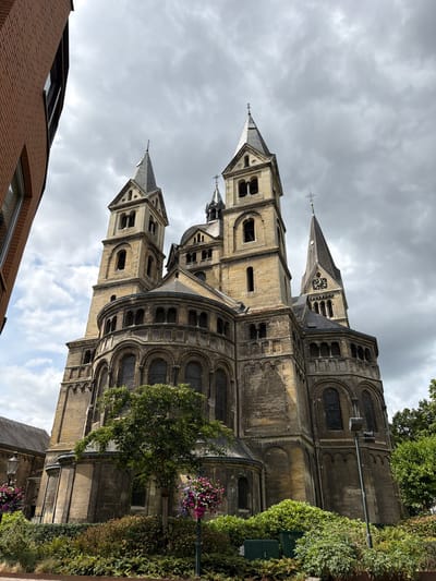 The image shows the exterior of the Munsterkerk, a Roman Catholic church located in Roermond, Netherlands. The church is constructed of stone and features multiple towers, a central dome, and arched windows. The building is surrounded by greenery, including trees and bushes, and there are hanging flower baskets on lampposts in the foreground. The sky is overcast. The perspective is from a low angle looking up at the church. There are no people or animals visible in the image.