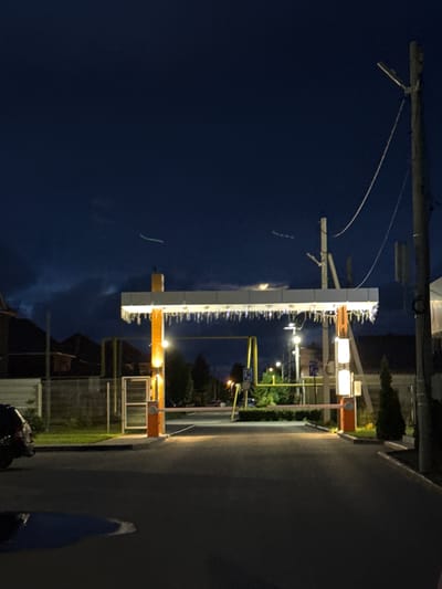 The image depicts a nighttime scene at what appears to be an entrance to a residential area or parking lot in Primorsky, Russia. A barrier gate system is prominently featured in the center, with an orange vertical post supporting a horizontal boom that is currently open. The gate structure is illuminated by lights, and decorative icicle-like lights hang from the underside of the horizontal boom. To the left of the gate, a dark-colored vehicle is partially visible. The road surface is asphalt, and to the right, a utility pole with wires and a street lamp is present. The background shows trees, fences, and buildings, suggesting a suburban or developed area. The sky is dark blue with some faint cloud formations and a few streaks of light that could be from aircraft or distant lights. A puddle of water is visible on the road in the foreground, reflecting the ambient light.