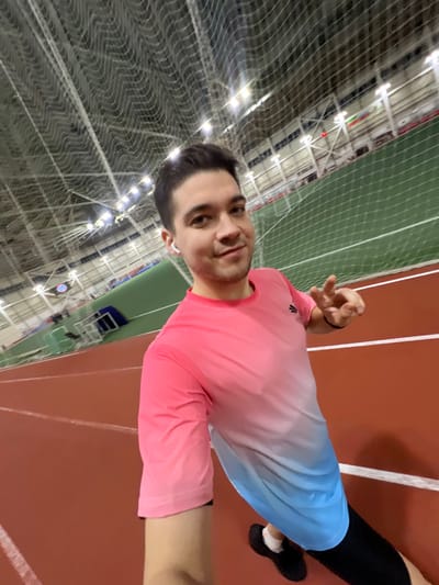 A person in a pink and blue shirt stands in a building with a green net ceiling. They are on a red running track, wearing black athletic bottoms and shoes. They are holding their hand up, making a hand gesture. The building has many lights.