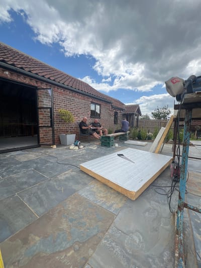 This is a daytime outdoor scene at a construction or renovation site located in Skipsea, United Kingdom. The foreground features a dark slate patio. A large, rectangular insulation panel or similar building material, light grey on top with a yellow-brown edge, lies horizontally on the patio. A handsaw with a black handle rests centrally on this panel. To the right, a blue metal scaffolding structure supports a platform on which a red and black Ryobi-branded power tool, likely a miter saw or chop saw, is positioned. An electrical cord extends from this area across the patio towards a circular saw or angle grinder resting near the base of a brick building.
In the mid-ground, three adult males are present. Two males are seated in black patio chairs to the left, appearing to be conversing or observing. A third male is seated on a makeshift bench composed of a wooden plank supported by two green plastic crates. All three individuals appear to be taking a break from work.
The background consists of a red-brick building with a dark, tiled roof. The building features a large, open doorway on the left, leading into an uncompleted interior space. Further to the right along the building, there is a dark-framed window and a smaller entrance with a tiled canopy. A small, potted tree in a grey square planter is positioned on the patio near the building. Beyond the immediate working area, a wooden fence and various green foliage are visible under a partly cloudy blue sky.