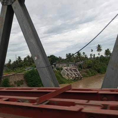 The image shows a view from a bridge, looking towards a rural landscape with a river. In the foreground, there are large, red metal beams of the bridge structure. In the mid-ground, a section of the riverbank is visible, with vegetation and a partially collapsed wooden structure. Further in the background, there are several houses and a dense forest of palm trees and other greenery. The sky is overcast with clouds. The scene appears to be in a rural area of Meuse, Indonesia, suggested by the tropical vegetation and the style of the houses. There are no people visible in the image.