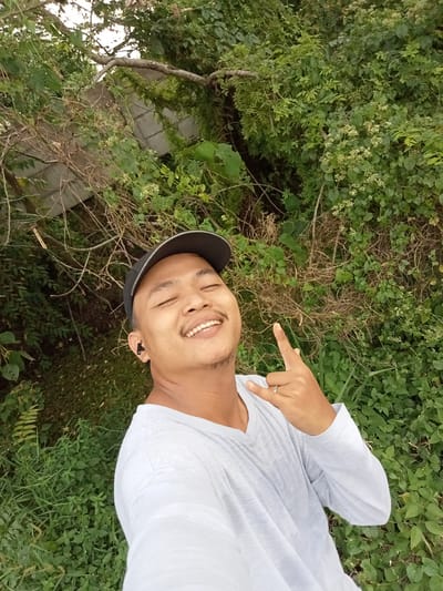 A selfie-style photograph captures a man in Teluk Kenidai, Indonesia, with a backdrop of lush green foliage. The man, wearing a white long-sleeved shirt and a dark baseball cap, smiles with his eyes closed and gestures a rock-and-roll sign with his right hand. He has an earbud in his left ear and a ring on his right ring finger. The environment appears to be outdoors, with a variety of plants and trees dominating the scene. Some man-made structures are partially visible in the upper left portion of the image, suggesting a semi-urban or semi-rural setting. The lighting indicates it is daytime.