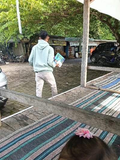 The image depicts an outdoor scene in Gembong, Indonesia, from a slightly elevated perspective. In the immediate foreground, the top of a dark-haired head with a pink flower hair clip is visible in the lower right. A multi-colored striped mat, featuring hues of blue, black, white, and orange, lies on a wooden platform. A prominent horizontal wooden beam separates this platform from the lower paved area.
A male individual stands in the mid-ground, facing away from the viewer, wearing a light mint green hooded sweatshirt and light beige trousers. He holds a blue folder or book and a white paper in his left hand, looking towards a row of shops. The ground beneath him and extending into the background is a wet, dark grey paved surface, likely interlocking pavers or cobblestones.
In the background, a series of rustic shops or stalls are arranged along the paved area, featuring corrugated metal roofs and wooden construction. Visible signs include "MANDIRI" and "BARBERSHOP," along with Indonesian text. Piles of coconuts and other produce are displayed outside these establishments. A silver vehicle's front fender is partially visible on the far left, near parked motorcycles. Towards the right, a black multi-purpose vehicle (MPV) is parked, with two motorcycles adjacent to it. A person wearing an orange garment is faintly discernible near the black MPV. Lush green tree foliage and dense vegetation dominate the upper background, indicating a verdant environment. A white canopy or tarp partially extends from the upper right, supported by a wooden post.