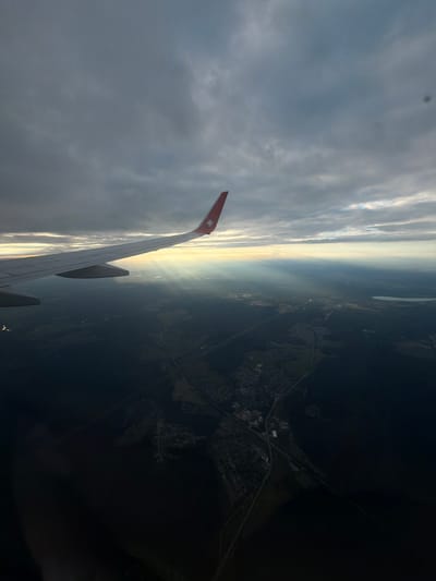 An aerial view captures an airplane wing extending from the left side of the frame, pointing towards a horizon where sunlight breaks through dramatic, dark clouds. The wing, predominantly white with a red tip featuring a star logo, casts a shadow over a landscape below. This landscape is a mixture of dark forest and scattered settlements, with faint roads visible. The sunlight creates dramatic rays piercing through the cloud cover, illuminating a section of the ground and a body of water to the right. The scene suggests the airplane is in flight over the area of Dubrovo (Uleiminskoe S/P), Russia, given the specific terrain features and likely position. The overall atmosphere is one of transition, with the dynamic interplay of light and shadow over the Russian landscape.