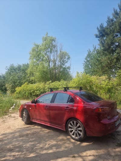 A red Mitsubishi Lancer sedan is parked on a dirt road in a rural area, possibly in Szczaki, Poland, on a sunny day. The car has a roof rack and is angled towards the left side of the frame. Lush green trees and bushes surround the road, with a clear blue sky overhead. The ground is sandy and dry, with shadows cast by the trees. There are no people visible in the image.