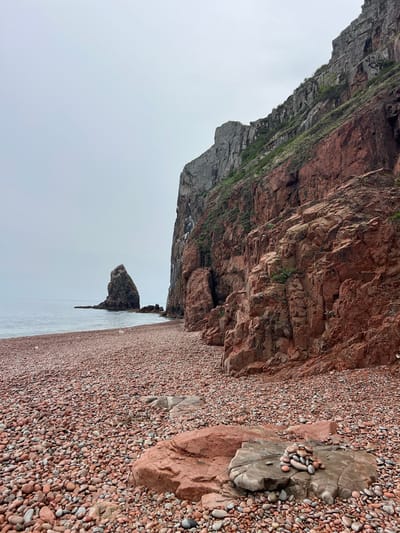 The image depicts a rocky beach in Russkiy, Russia. A large red rock cliff dominates the top of the frame. Vegetation clings to the cliff face. To the right of the cliff is a beach covered with small, red pebbles. A large grey boulder is on the beach with a collection of smaller stones stacked upon it. The sea is visible in the lower part of the image, with a rock formation jutting out of the water. The sky is overcast.