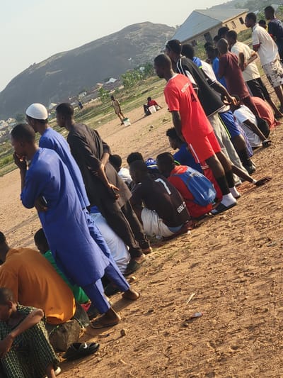 A group of approximately 15 men are lying prone on a sandy, open area in what appears to be Ibo, Nigeria. They are arranged in a line, with some individuals wearing traditional Muslim clothing such as a blue robe and white kufi cap. Others are in casual wear including t-shirts and shorts, with one wearing a red jersey with "ROYSTARS" printed on the back. They are lying face down, possibly engaged in prayer or some other form of communal activity. Behind them, a hilly landscape is visible, along with buildings and vegetation. The ground is mostly bare, sandy soil. The angle of the photo suggests it was taken from a slightly elevated position.