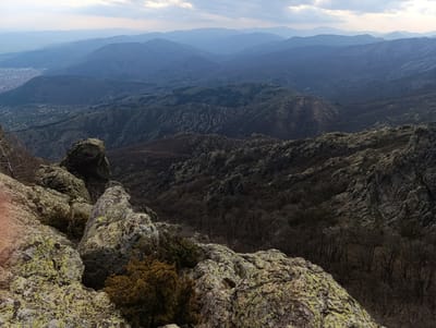 The image is a high-angle landscape view from a rocky, mountainous terrain, looking toward a cityscape in the distance. The foreground consists of large, moss-covered rocks with sparse vegetation. A city is visible in the distance, and layered mountains recede into the background under a cloudy sky. The lighting is diffused. The time of day appears to be either early morning or late afternoon.