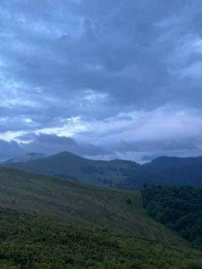 This is a vertical, medium-wide shot of rolling hills and mountains under a cloudy, twilight sky. The foreground is a steep slope covered in low-lying green shrubs. A path is visible winding up the slope. In the mid-ground, two large, rounded hills dominate the scene, with the left hill appearing slightly higher and shrouded in mist at its peak. The right mid-ground is a densely forested slope. The background consists of more distant, darker, forested mountains. The sky is filled with heavy, dark blue and gray clouds, with hints of lighter sky peeking through in the upper center. The overall lighting suggests it is either dawn or dusk. The location is the area of Ust'-Chorna, Ukraine. There are no people or animals visible in the image.