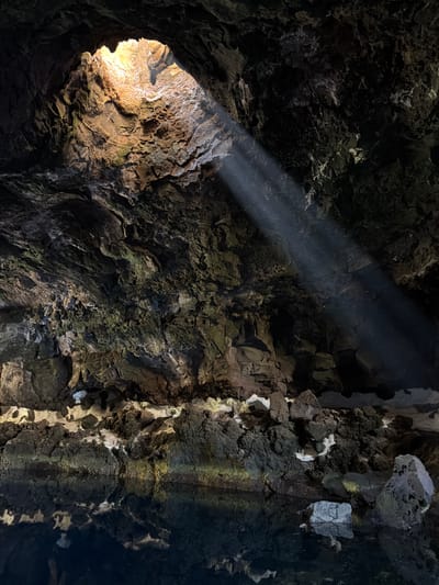 The image shows a view inside Cueva de los Verdes, a lava tube located in Haría, Spain. Light enters the cave through an opening at the left, illuminating the rough rock walls and creating a beam of light. The walls are composed of dark volcanic rock with patches of lighter-colored mineral deposits. The structure of the rock suggests a cave environment.