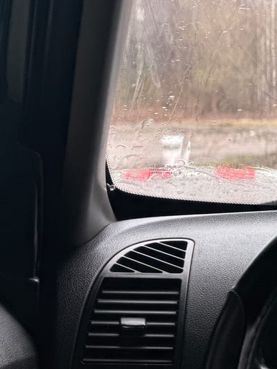 This close-up image, taken from inside a vehicle, captures a wet car window and a section of the dashboard. The left side of the frame is dominated by the car's window, which is heavily streaked and dotted with water droplets, suggesting either rainfall or a car wash. Through the blurred, wet glass, a muted, indistinct outdoor environment is visible, with a faint red hue reflecting or appearing in the distance.
Along the edge of the window, a black frit band is discernible, and mirrored text indicating the glass manufacturer and safety standards is visible. The text, viewed from inside, reads "LAMINATED," "SAINT-GOBAIN SEKURIT," "E2," and "43R-000105." This implies the window is European-standard laminated safety glass.
The right side of the image showcases a dark, textured plastic surface of the car's dashboard. A vertical air conditioning or heating vent with multiple slats is prominently featured in the lower right quadrant. The overall lighting suggests an overcast day or an interior shaded from direct sunlight, with the exterior appearing brighter but diffused. No people are visible, and the scene implies the vehicle is either stationary during precipitation or undergoing a wash in Klenovskoe, Russia. The focus on the water droplets and the interior details creates an intimate perspective from within the car.
