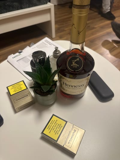 An overhead-angled photograph captures a static indoor scene on a white table surface in Höri, Switzerland. Positioned centrally is a nearly full bottle of Hennessy Very Special Cognac, its clear glass revealing an amber liquid, topped with a gold foil seal. To the left of the cognac bottle, a small succulent plant in a light grey cylindrical pot is visible. Further left, a small, dark-capped bottle, possibly perfume or aftershave, sits next to a small clear glass bottle with a silver pump dispenser. Partially obscured behind these items is a stack of white papers, with the word "sanitas" discernible on the top sheet. To the right of the cognac bottle, a black rectangular object, possibly a phone or glasses case, rests flat. In the foreground, a pack of Dunhill Classic Gold cigarettes lies flat, displaying a yellow warning label with text in multiple languages. Another identical pack of Dunhill Classic Gold cigarettes stands upright behind the succulent. The background reveals a wooden floor, a white furniture frame with a dark grey textile on it, and the lower legs and feet of an individual wearing dark trousers and shoes. No direct human interaction with the items is observed.