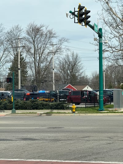 The image depicts an outdoor scene at the intersection of Stoney Creek Road and Georgesville Road in Columbus, Ohio, United States. The time is daytime under an overcast sky, with bare deciduous trees indicating a non-foliage season.
In the foreground, an asphalt roadway is visible, marked with a white stop line and a section of reddish paving. A concrete curb separates the road from a median area featuring green grass and shrubbery.
Behind the curb and shrubbery, a black metal fence runs horizontally across the midground. Several vehicles are situated behind this fence. At least two dark-colored Chevrolet Tahoe-style SUVs, marked with "SHERIFF" in yellow lettering, are parked. One sheriff's vehicle displays active flashing blue and red emergency lights on its roof and possibly in the rear window. The driver's side front door of this vehicle appears slightly ajar. Adjacent to the law enforcement vehicles, a dark red minivan is parked with its driver's side sliding door and rear hatch open, revealing items inside. A yellow fire hydrant is visible behind the fence, in front of the minivan.
On the left, a shorter green pole holds a red pedestrian traffic signal. On the right, a tall, prominent green traffic light pole supports multiple dark, unlit signal heads and a green street sign reading "STONEY CREEK RD". Various sensors and hardware are attached to this pole. A gray utility box is positioned on the far right, behind the fence.
The background consists of numerous bare deciduous trees and some evergreen trees. Overhead utility poles and power lines are visible. Beyond the vehicles, residential-style buildings can be seen, including one with a red roof and another with a white facade and peaked roof. No individuals are clearly visible in the frame.