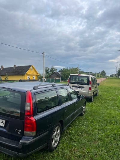 The image captures a rural scene in Бучацінскі сельскі Савет, Belarus. In the foreground, a dark blue Volvo V70 station wagon is parked on the left side of a gravel road. The car is facing away from the camera, with its rear visible. To the right of the Volvo, a silver minivan, possibly a Fiat Doblo, is also parked on the grass verge, facing the same direction. The minivan's license plate reads "7526 BX-5 BY".
A bright yellow house with a dark roof and white trim is situated behind the Volvo on the left side of the road. Further down the road, more houses and trees can be seen. The sky is overcast with grey clouds, suggesting a potentially gloomy or rainy day. The road itself appears to be unpaved, made of gravel or dirt, and it curves to the right as it recedes into the distance. Utility poles with wires are visible along the side of the road. The area is covered in lush green grass, indicating a rural or semi-rural setting.