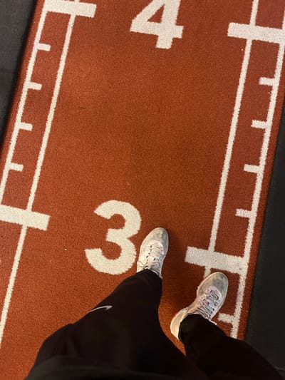 An overhead photograph depicts the lower body of an individual standing on a red-orange indoor athletic track surface within a facility in Clermont-Ferrand, France. White painted markings on the track include the number "3" beneath the subject's feet, and the upper segment of the number "4" visible further up the track. Parallel white lane lines and perpendicular white hash marks, indicating measurement intervals, are also present. The subject is wearing black athletic trousers, which feature a white Nike swoosh logo on the outer left thigh. Both feet are shod in light grey or off-white athletic shoes with white laces and minor orange or pink accents; these shoes exhibit some surface dust or wear. The track surface appears to transition to a dark, solid-colored floor or wall at its edges. The individual is stationary, facing forward from the perspective of the camera.