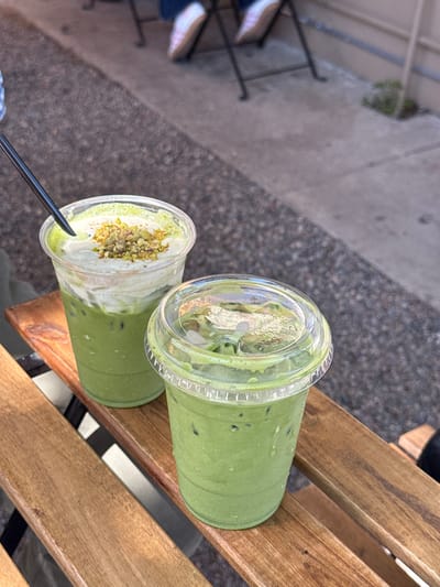 Two plastic cups of green matcha drinks are on a wooden outdoor table in Chula Vista, United States of America. The cup on the right has ice and a clear plastic lid. The other cup has a black straw and whipped topping covered with chopped nuts.