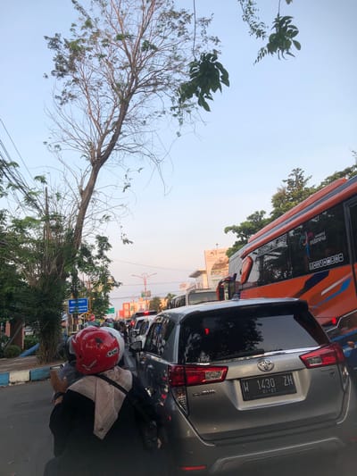 This media file captures a traffic jam on a street, likely in Sidoarjo, East Java, as suggested by the text on the vehicle's license plate and surrounding signs. The scene is dominated by vehicles, including a large orange bus with "wonderful indonesia" branding and a silver car with license plate "W 1430 ZH" and an expiry date of "01-27". A person wearing a red helmet and a light-colored scarf is visible on a motorcycle in the foreground, indicating the activity is commuting or traveling. The presence of traffic lights and the vehicles' brake lights suggest a standstill or slow-moving traffic. The time of day appears to be late afternoon or early evening, with soft, warm lighting from the setting sun casting long shadows. The sky is clear, suggesting fair weather. The environment includes trees and greenery alongside the road, and buildings and billboards in the background, typical of an urban or suburban setting. The overall mood is one of everyday life, with the common experience of traffic congestion. Text visible includes Indonesian words like "BERENTI" (stop) and place names such as "prigen" and "bundu", further grounding the location.