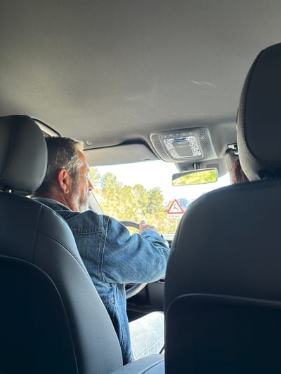 The image depicts the interior of a vehicle, likely a car or van, with a man reclined in the driver's seat. The man, who appears to be middle-aged, has graying hair and is wearing a denim shirt. He is leaning back in the seat, with his head resting on the headrest. His hands are visible on the steering wheel.
The vehicle's interior features a gray headliner and black leather seats. A rearview mirror and a panel with illuminated buttons are visible near the top of the frame.
Outside the vehicle, a glimpse of the surroundings is visible through the windshield, revealing trees and a road sign indicating a traffic island and merge, indicating that the vehicle is in transit.
The road sign visible through the windshield of the car has the letters “CV-750” written on it, indicating the car is on the CV-750 road in Benissa, Spain.