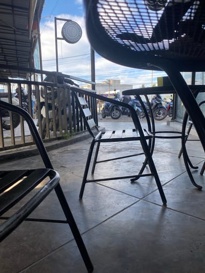 The image shows an outdoor seating area with metal chairs and tables. In the background, several motorcycles are parked behind a metal fence. Above the fence, a sign that reads "DOCTOR PENY CLINIC" with a silhouette of a face is visible. The sky is cloudy with patches of blue. The ground is paved with concrete tiles. The location is identified as Keudeublang, Indonesia.