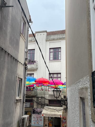 This is a medium-angle, full-shot photograph of a narrow street in Águeda, Portugal, on a cloudy day. The foreground is dominated by the textured gray facade of a building on the left and the rough stucco of a building on the right. Black electrical wires crisscross the scene, obscuring parts of the background. Suspended across the street are numerous colorful umbrellas, predominantly in shades of blue, pink, red, yellow, and green, creating a vibrant canopy overhead. These umbrellas are attached to their hooks and are open, offering a decorative effect.
Below the umbrellas, on the left side of the street, a portion of a building shows windows with purple frames. A sign with the word "RIA" repeated vertically is visible. Further down, an air conditioning unit is mounted on the wall of the building. On the right side, a vintage-style street lamp with a black metal ornate bracket is attached to the wall. Below the street lamp, signs for a shop, one reading "Salão de Chá · Doces Regionais," are visible.
In the background, a white building with distinctive vertical architectural elements and purple-framed windows stands out. A balcony with a curved railing is visible on this building. The roofs of buildings, covered in red tiles, are also visible against the overcast sky. The scene captures a characteristic decoration found in Águeda, known for its "Umbrella Sky Project" which decorates its streets with colorful umbrellas, particularly during the Águeda Art Festival.