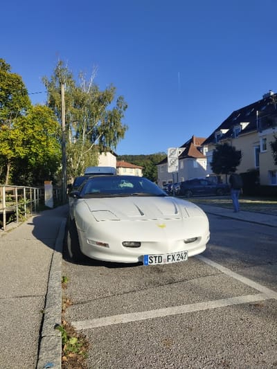 The image displays a white 4th generation Pontiac Firebird Trans Am convertible, equipped with a black soft-top roof and a white roof box, parked head-on in a marked asphalt parking space in Herrsching am Ammersee, Germany. The vehicle exhibits a yellow bird emblem on its hood and displays the German license plate D STD FX 247. To the left, a grey paved sidewalk borders a stone curb, beyond which a metal railing is partially visible. The immediate environment includes sparse vegetation between the curb and pavement. In the background, several light-colored residential buildings with brown roofs line the street. A dark blue car is parked further down the road, partially obscuring a pedestrian wearing a dark jacket and light trousers walking away from the camera on the right-hand sidewalk. Two white circular traffic signs are visible near the buildings. Green foliage and a tall deciduous tree frame the left side of the scene, with a tree-covered hill in the distant background. The sky is clear blue, traversed by a faint white contrail. The scene is brightly lit by natural sunlight, casting distinct shadows.