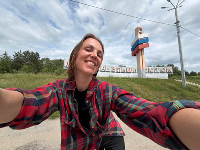 A woman with long brown hair is taking a selfie outdoors. She is wearing a red, blue, and yellow plaid shirt over a black top. Behind her, a sign with Cyrillic lettering reads "МА-ВЛАДИВОСТОК". A pole with three colored stripes (white, blue, and red) stands to the right of the sign, topped by a coat of arms. A lamppost is also visible to the far right. The background consists of a grassy slope, trees, and a cloudy sky. The ground in the foreground is a gravel path.