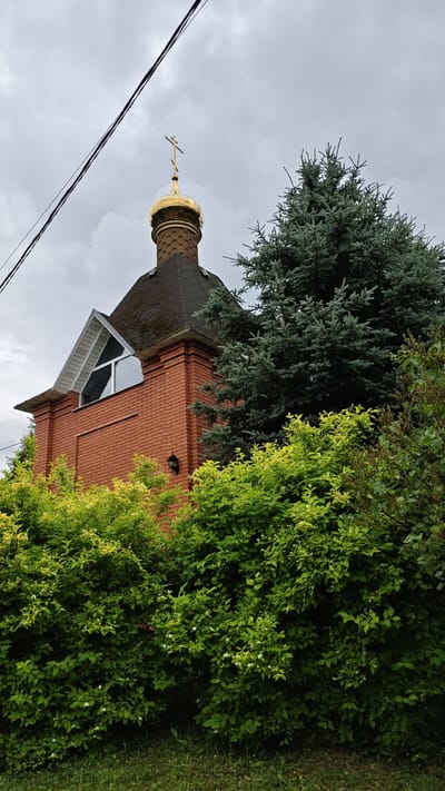 This media file captures a picturesque scene in Sof'ino, Russia, under a cloudy sky. The primary subject is a small, red-brick Orthodox church, its golden dome and cross glinting even in the overcast light. The church is partially obscured by lush greenery, with a mature, blue-green conifer tree standing tall beside it, and a dense bush with bright green leaves and small white flowers in the foreground. A large, triangular window with white framing is visible on the side of the church, reflecting the sky. The roof of the church appears to be dark, possibly slate or shingles, with some moss or discoloration evident. Electrical wires are visible stretching across the top of the frame, suggesting the presence of nearby infrastructure. The overall mood is peaceful and serene, with the natural elements harmonizing with the religious structure. The time of day is indeterminate due to the cloud cover, but the soft, diffused light suggests it is daytime, possibly mid-morning or afternoon. There is no visible text in the image.