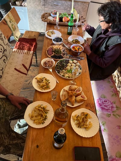 In Sovetskaya Gavan', Russia, a woman with dark, curly hair, wearing glasses and a dark purple hoodie with a vest, is seated at a wooden table laden with food. She is using a fork to pick up food from a white plate. The table is covered with various dishes, including plates of french fries, a charcuterie board with meats and cheeses, a bowl of olives, and a plate with pastries wrapped around hot dogs. There are also bottles of liquid, a wine glass, and a small glass of what appears to be vodka. In the background, a wooden bench is visible, draped with a patterned textile and a red blanket. The flooring is made of rough wooden planks. The scene is illuminated by natural light, creating a warm and inviting atmosphere.