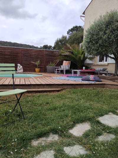The image depicts a backyard in Saint-Paul-et-Valmalle, France. A wooden deck with a small pool is surrounded by a grassy lawn with stone pavers. The deck features various objects, including a statue, potted plants, cushions, and furniture, suggesting a relaxation or leisure area. A wooden fence is visible in the background, with trees beyond. To the right, part of a building is visible. The scene is captured on an overcast day. No people are present.