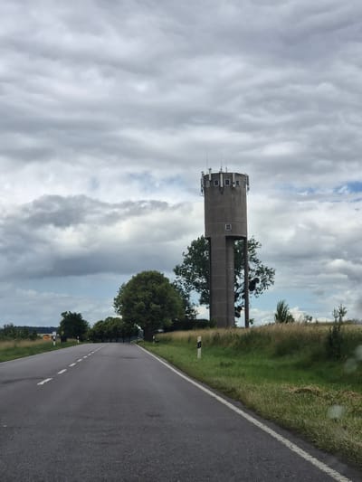 An outdoor, eye-level, medium shot captures a concrete water tower with antennas on top, positioned to the right of a paved, two-lane road. The road curves into the distance on the left, flanked by grassy fields and trees. The sky is overcast with heavy gray clouds, though patches of blue are visible. The water tower is cylindrical with a square opening and a circular top with several antennas. Several trees, some dense and green, others more sparse, surround the tower. The road has a dashed white line in the center and a solid white line on the right edge. There are no people or vehicles visible in the frame. The scene appears to be in a rural setting, potentially near the city of Schengen, Luxembourg, based on the architecture and landscape.