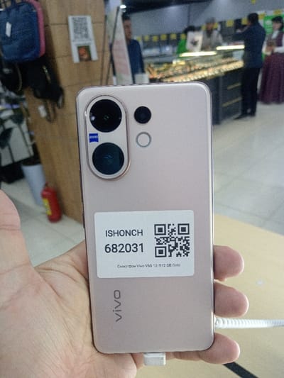 A light-skinned human hand holds a gold-colored Vivo V60 smartphone, 12GB RAM/512GB storage variant, in an indoor retail environment located in Shofirkon, Uzbekistan. The smartphone's back features a prominent camera module in the upper left, containing two vertically aligned large circular lenses—the upper one marked with a blue "ZEISS" logo—a smaller circular lens to the right, and a flash/sensor below it. A white rectangular sticker is affixed to the lower half of the phone's back, displaying "ISHONCH," the number "682031," a QR code, and the text "Смартфон Vivo V60 12/512 GB Gold." The "vivo" brand logo is vertically printed near the bottom of the phone. A white security tether connector is visible at the phone's bottom edge, attached to a coiled white cable. The blurred background reveals a store interior: a wooden display stand with a dark blue backpack and a visible QR code on the left, and a red fire extinguisher on the floor. In the central and right background, illuminated glass display cases exhibit merchandise. Three individuals are partially visible in the background: one person in a dark blue top behind the left display, another in a dark jacket observing items at a central display, and a third person in a dark top and long maroon skirt on the far right. No direct interaction between the background individuals or with the person holding the phone is evident.