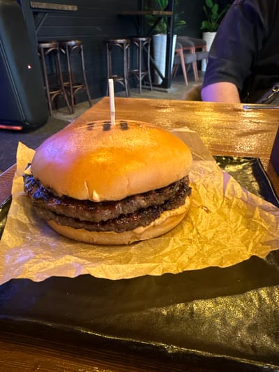 This media file features a close-up, overhead shot of a double cheeseburger, presented on a bed of crumpled brown paper. The burger has a golden-brown bun, layered with two thick, grilled beef patties. A white toothpick is inserted through the top bun. The burger is placed on a dark, possibly leather or vinyl surface. To the left of the burger, a person's arm, wearing a dark jacket, is partially visible. The background shows glimpses of what appears to be restaurant seating with wooden chairs and tables, suggesting an indoor dining environment. The lighting is somewhat dim, with warm tones reflecting off surfaces, hinting at an evening or indoor setting. No visible text is present in the image. The overall impression is that of a casual dining experience, focused on the food. Given the provided location context of Manzherok, Russia, this is likely a meal being enjoyed at a restaurant or cafe in that area.