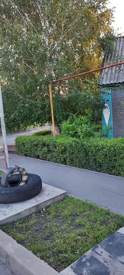 The image depicts an outdoor scene in Shakhty City District, Russia. In the foreground, two black tires are stacked on a concrete slab. A paved pathway runs alongside a neatly trimmed green hedge. Behind the hedge, a tree with white bark and green leaves is visible, with its branches reaching towards a rustic building. The building has a grey, corrugated roof and a blue-painted section adorned with a white diamond shape. A rusty orange pipe extends horizontally across the scene, supported by a yellow post. The ground is partially covered with sparse green grass and dark soil. The lighting suggests it is late afternoon or early evening.