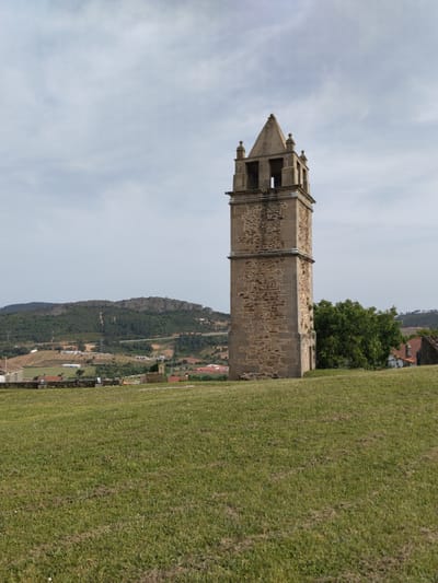 The image shows a tall, stone bell tower located in Mogadouro, Portugal. The tower is the main subject and is composed of two primary sections, the lower one being wider and constructed with rough-hewn stones, while the upper section is narrower with smooth stone corners and features open archways at the top, topped with a pyramidal roof. The tower stands on a grassy hill that slopes down towards the foreground. Behind the tower, there are trees and rooftops that suggest a small village. In the background, rolling hills and mountains covered in forests are visible under a cloudy sky.