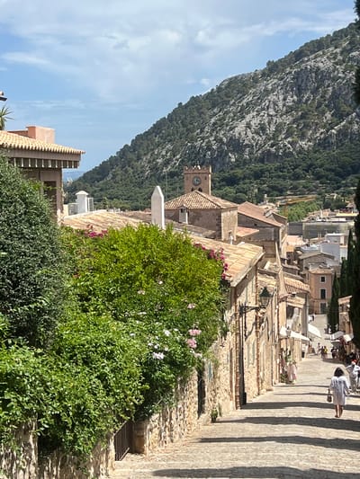 The image depicts a sun-drenched, cobblestone street in the town of Pollença, Spain, leading uphill towards a densely forested mountainside. The architecture is consistent with Mediterranean style, featuring terracotta-tiled roofs and stone buildings. In the foreground, lush green foliage and flowering bushes line the left side of the street, cascading over a stone wall. A dark metal gate is visible within the wall.

Further up the street, several people are visible walking and browsing shops. The buildings on either side of the street have awnings and display merchandise. A prominent feature in the mid-ground is a clock tower with a square base and a pyramidal roof, characteristic of a church or town hall. In the distance, the steep, rocky mountain slopes are covered in dense green trees, suggesting a natural landscape bordering the town. The sky is a clear blue with a few scattered clouds, indicating fair weather. The overall scene conveys a sense of a charming, historic European town on a bright, warm day.