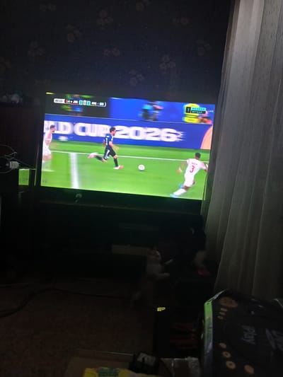 The image shows a television screen displaying a soccer match. The score shown is Jordan 0, Argentina 2. The time on the clock is 49:23. The text "WORLD CUP 2026" is visible across the bottom of the screen. One player in a blue uniform is running with the ball, and another player in a red and white uniform is running towards him. The environment appears to be a dimly lit room, with a curtain visible on the right side of the image. There is no information in the image to suggest the location is Mishkovo-Pohorilove, Ukraine.