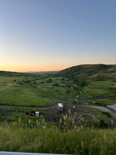 A landscape photograph, rotated 90 degrees, depicts a rural scene. The sky, transitioning from blue to orange, occupies the left portion of the frame. A hill covered in dense green vegetation runs vertically. A dirt road follows the side of the hill. A red and white vehicle rests near the bottom of the image. A small structure and scattered white objects are visible along the road. Based on the scene the location is likely Vratitsa, Bulgaria.
