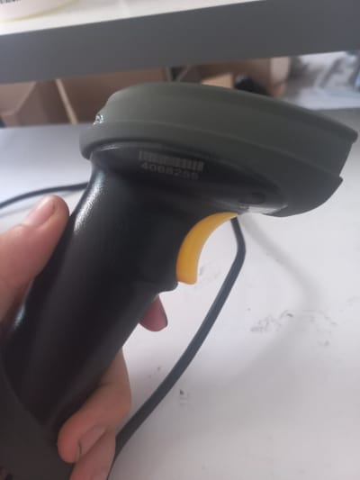 The image shows a close-up of a black handheld barcode scanner with a yellow trigger, held by a person's hand. The scanner has a green top section. A barcode and the number "4068255" are visible on the scanner. The background is blurry, showing a white surface and some shelves or boxes. The image does not contain any information related to the city of Yorkatay, Uzbekistan.