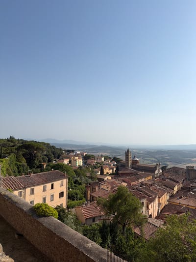 The image shows an aerial view of Massa Marittima, Italy, on a clear, sunny day. The foreground is dominated by a stone wall, part of the city's fortifications, with greenery growing on its top. Beyond the wall, the terracotta-tiled roofs of buildings characteristic of Italian hill towns spread across the landscape.
In the mid-ground, several buildings are visible, including a prominent yellow structure with multiple windows. A cluster of trees and dense foliage fills the spaces between the buildings, adding a lush, green element to the scene.
Further in the background, the distinctive bell tower of the Cathedral of San Cerbone rises above the surrounding architecture, accompanied by the main body of the cathedral with its striped facade and arched windows. More buildings, in various colors like pink and yellow, are interspersed with trees.
The cityscape extends towards a valley with rolling hills and agricultural fields, characteristic of the Tuscan countryside. The distant landscape fades into a hazy blue, with faint outlines of mountains on the horizon. The sky is a clear, pale blue, devoid of clouds. No people are visible in the image.