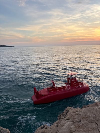 A red submarine-like vessel with a canopy is seen in the turquoise waters of the Adriatic Sea near Primošten, Croatia. The vessel is observed from a rocky shoreline, with a person visible in the pilot's seat. The sky above is a gradient of pale blue and orange, indicating either sunrise or sunset, with some wispy clouds. The sea has gentle waves, and a small island is visible in the distance.