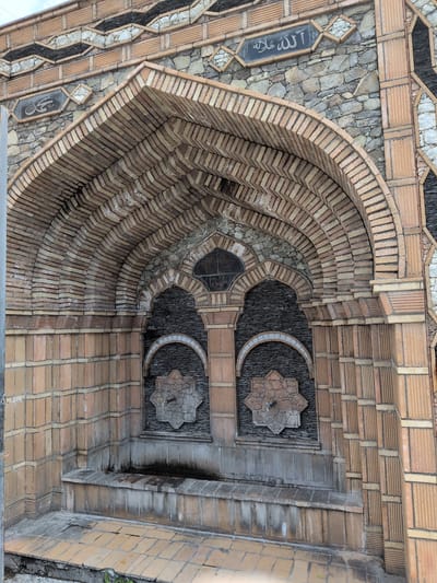 The image shows a close-up view of an ornate, historic fountain structure made of brick and stone, located in the city of Şəki, Azerbaijan. The fountain features an arched brick facade with intricate geometric patterns. Two arched niches are visible within the main arch, each containing a star-shaped decorative element and a water faucet. Below the niches, a stone bench serves as the basin of the fountain. The stonework above the brick arch displays Arabic calligraphy. The overall structure appears weathered but well-maintained, suggesting it is a significant cultural landmark. The paving in front of the fountain is also made of stone tiles.