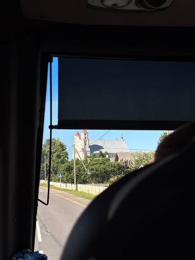 The image, taken from inside a vehicle, captures a view through a window with a partially drawn roller blind. The foreground is dominated by the dark interior of the vehicle, including a curved, dark surface on the right side which appears to be a headrest or seat. The window looks out onto a rural scene in Васілішкаўскі сельскі Савет, Belarus.
In the middle ground, a paved road runs diagonally from the lower left to the middle right of the frame. On the left side of the road, a white pedestrian crossing symbol is visible on the asphalt. To the right of the road, a low, ornate concrete fence lines the property. Beyond the fence, lush green trees and shrubs are abundant. A blue pedestrian crossing sign on a pole is situated amongst the trees.
In the background, a prominent church with a light-colored facade and red-tiled roofs stands out against a clear blue sky. The church features multiple towers and a cross atop its central spire. The architectural style suggests a historic European building. The area around the church is characterized by more trees and some residential-looking buildings with tiled roofs. The overall impression is of a sunny day in a peaceful, possibly historic, rural setting in Belarus.