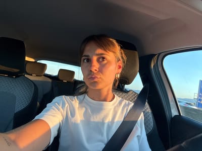 A woman with brown hair and brown eyes is in the driver's seat of a car, looking at the camera. She is wearing a white t-shirt and a seatbelt. The sun is setting, casting a warm glow on her face and arm. The car appears to be on a highway. This scene is consistent with a location in the city of Reventin-Vaugris, France, given its geographical location and the typical presence of highways for travel.