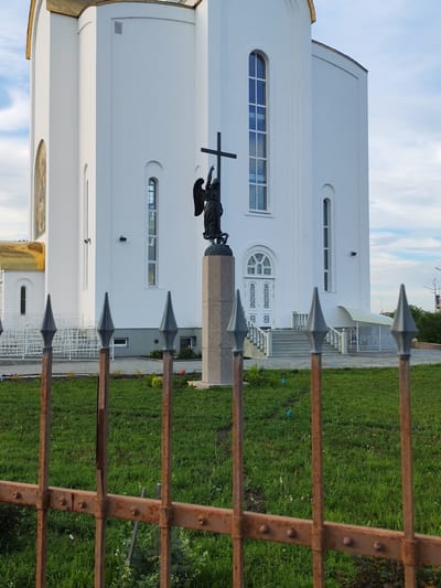 The image shows a white church with a golden dome on the left. In front of the church, there is a statue of an angel holding a cross. The statue is on a stone pedestal. There is a rusty metal fence in the foreground. The scene appears to be in an urban area, possibly a park or public space. The sky is partly cloudy. The image was taken in Kurgan, Russia.