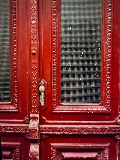 The image displays a close-up view of a deep red wooden double door located in Arthès, France. The door features two vertical rectangular panels of gridded or wired glass, one on the left and one on the right, both showing scattered white residue or dirt. The wooden frame around these glass panels, and along the central vertical stile separating the door sections, is intricately carved with repeating floral and geometric patterns. A decorative metallic doorknob, appearing gold-toned with silver-toned accents, is affixed to the right side of the central stile. The lower sections of the door also exhibit carved wooden panels. The entire structure shows signs of wear, including minor scratches and faded areas on the paint, indicating age and exposure to elements. No individuals are present, nor are any actions or interactions depicted.