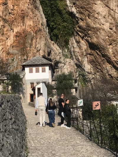 The scene depicts a paved stone pathway leading towards the Blagaj Tekija (Dervish Monastery) in Blagaj, Bosnia and Herzegovina. The pathway, constructed of irregular stone pavers, is bordered on the left by a stacked stone retaining wall and on the right by a black metal railing and sparse vegetation. In the background, a multi-story, white-washed traditional building with a grey stone roof and multiple rectangular wooden-framed windows is visible, built directly into the base of a towering, rugged, brown and grey limestone cliff face. Green foliage grows in crevices of the cliff.
Four individuals are present on the pathway, walking away from the viewer towards the monastery. From left to right:
1. A male in a light-colored vest over a white shirt and light-colored trousers, carrying a dark object in his left hand.
2. A female with dark hair, wearing a dark top and blue jeans.
3. A male with a bald head, wearing a dark shirt and dark trousers, holding a white object in his right hand.
4. A female with dark hair, wearing a dark jacket, dark trousers, and white sneakers.
To the right of the pathway, beyond the railing, two square signs featuring stylized Arabic script are visible; one is light green, and the other is reddish-brown. A black ornamental street light stands on the left side of the pathway, near the stone wall.
