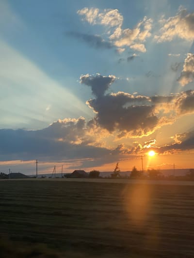 The image depicts a rural landscape at sunset, with a field in the foreground and a sky filled with dramatic clouds and sun rays. The field appears to be recently mown, with distinct parallel lines of cut grass. In the background, there are a few scattered houses and utility poles, suggesting a sparsely populated area. The sun is low on the horizon, casting a warm, golden light that illuminates the clouds and creates a bright glare. The sky transitions from a clear blue at the top to shades of orange and yellow near the sun. The overall impression is one of natural beauty and tranquility. The image is likely taken in or near the city of Svetlîi, Moldova, given the rural setting.