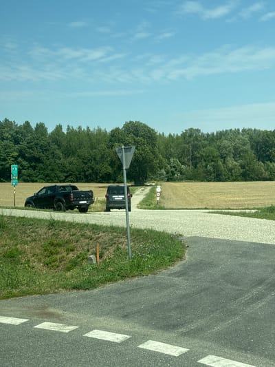 The image depicts an outdoor scene with two black vehicles parked on a gravel surface near a road. In the foreground, a portion of an asphalt road with white dashed lines is visible. To the left, a green signpost displays various arrows and symbols. Behind the vehicles, a field of harvested golden crops stretches into the distance, bordered by a dense line of green trees under a bright blue sky with scattered white clouds. A grey traffic signpost with a triangular top is positioned to the right of the gravel area. A smaller sign with a red circle and a bicycle symbol is visible on a pole near the field. The landscape suggests a rural or semi-rural setting. It is not possible to definitively identify the city of Wolfsthal, Austria, from the visual information alone.