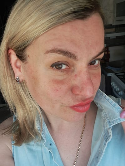 A headshot-style portrait displays an adult female in an indoor kitchen setting located in Yegoryevsk, Russia. The subject has fair skin, blonde hair, and brown eyes, with visible freckles and some minor redness on her face. She wears light eyeliner and coral-colored lipstick, directing a focused gaze towards the viewer with a slight pout. She is attired in a light blue, denim-style sleeveless top featuring a collar. Accessories include a thin gold-colored chain necklace and a silver-colored hoop earring with small red stones in her visible left ear. Her right hand, partially visible with a pink-painted fingernail, is positioned at the collar of her top. The background on the right side of the frame reveals a white gas stove with multiple control knobs, adjacent to dark counter-tops or shelving, and a light-colored cloth hanging.