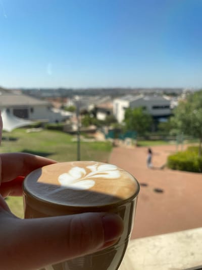 A hand holds a latte with latte art. The creamy surface is adorned with a delicate floral design. The background is a blurred urban landscape. Buildings, trees, and a pathway are visible, bathed in sunlight under a clear blue sky. The scene conveys a sense of relaxation and enjoyment of a coffee break with a scenic view.
